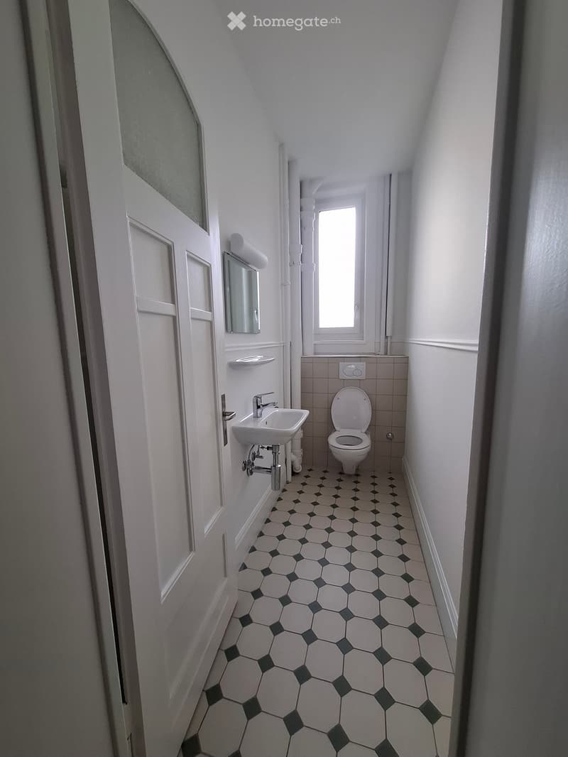 Small bathroom with hexagonal white and black tiles, a white sink with a chrome faucet, a toilet, a mirror, and a window letting in natural light.