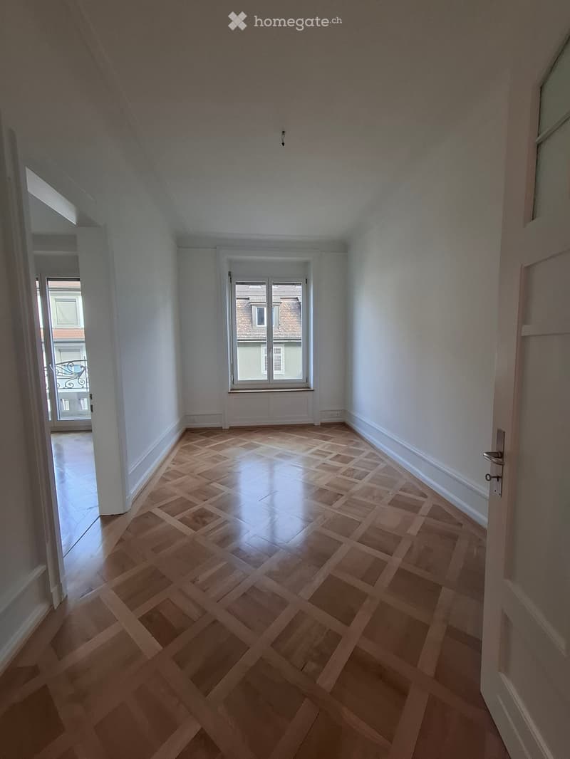 Empty room with wooden parquet flooring, white walls, and a window overlooking a building. No furniture or equipment present.