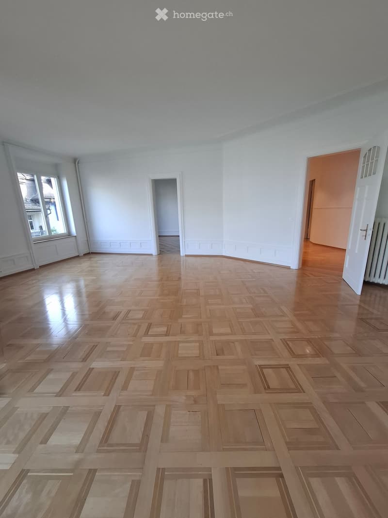 Empty room with light wooden parquet flooring, white walls, and large windows allowing natural light. The room features several doorways leading to adjacent spaces.
