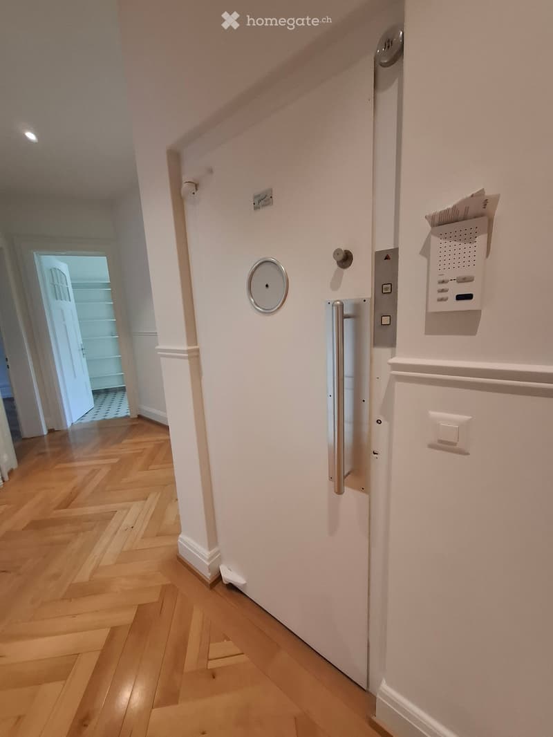 Hallway with wooden flooring, white walls, and a door with a security system and intercom.