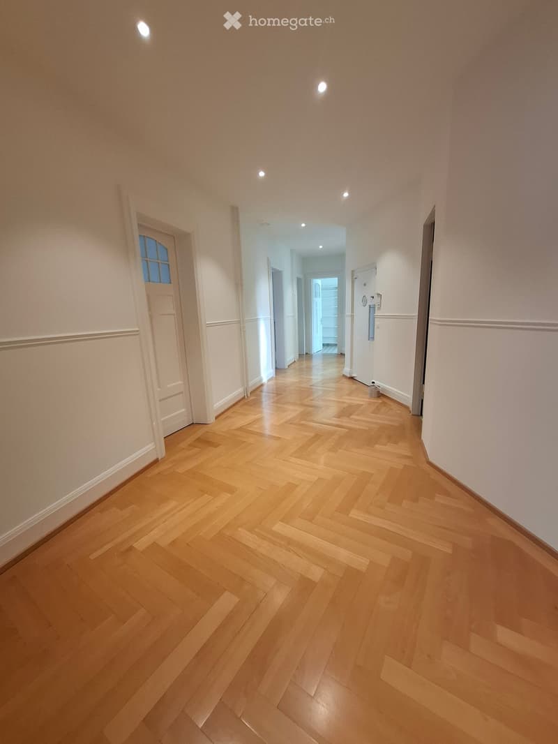 Empty hallway with wooden herringbone flooring, white walls, and recessed ceiling lights. Doors lead to adjacent rooms.