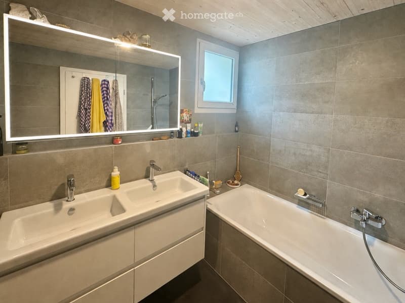 Modern bathroom with double sink, bathtub, large mirror with LED lighting, and various toiletries on the counter.