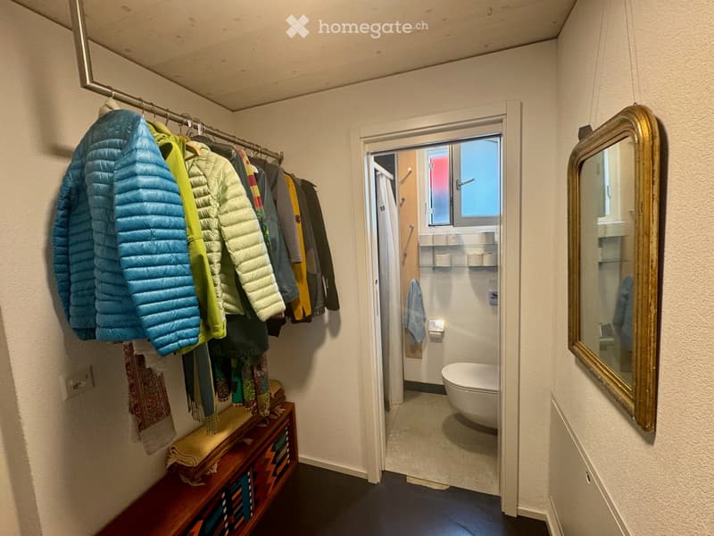 A narrow hallway with a coat rack holding jackets, a small wooden bench with folded towels, and an open door leading to a compact bathroom with a toilet, shelves, and a towel.