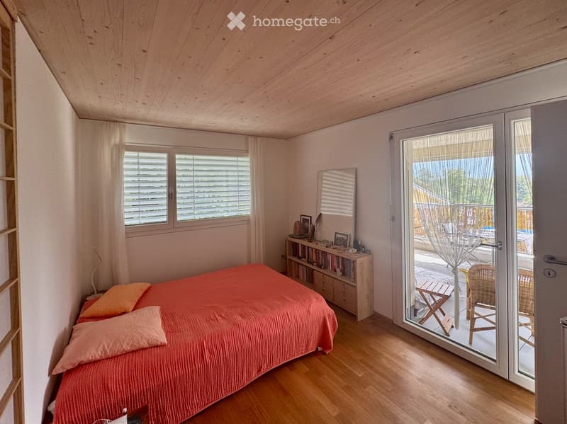 Bright bedroom with wooden floors, a large bed with orange bedding, a nightstand, a bookshelf, and a sliding glass door leading to a balcony with outdoor furniture.
