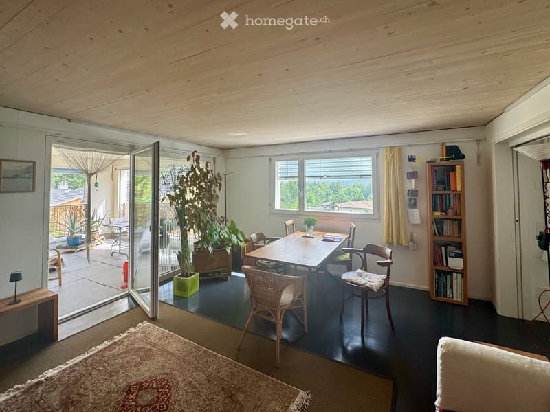 Bright, open room with wooden ceiling, large windows, and sliding glass doors leading to a patio. Features a sofa, dining table with chairs, bookshelf, and a large potted plant.
