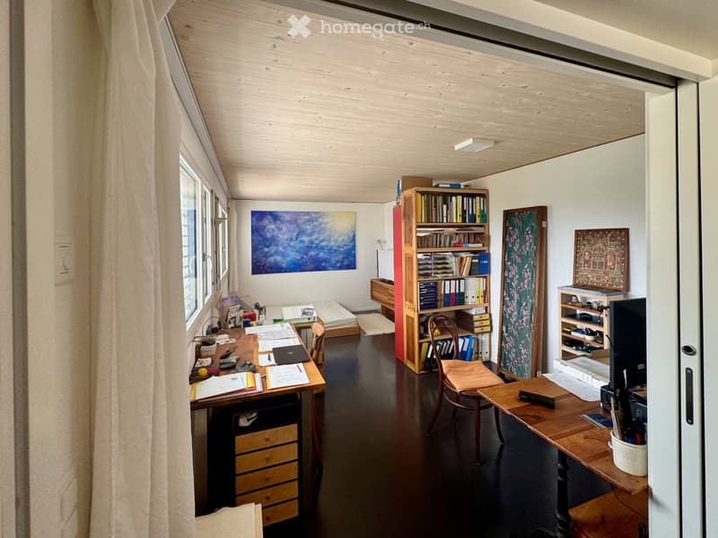 A cozy, well-lit room with wooden furniture, a large window with curtains, a desk with a laptop, a bookshelf, and a comfortable chair.