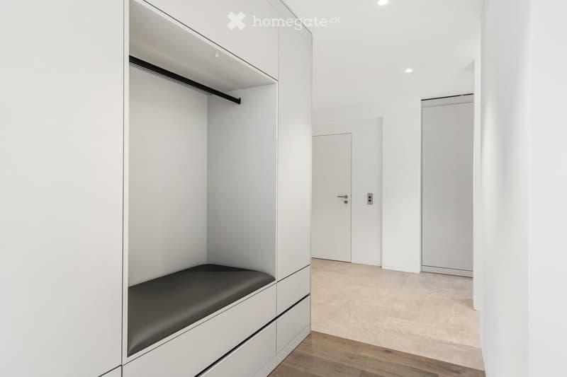 Modern, minimalist hallway with white walls, wooden flooring, built-in storage unit with black seating, and two doors.