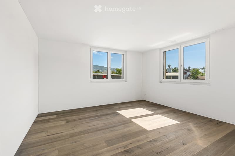 Empty room with white walls, wooden flooring, and large windows offering a scenic view.