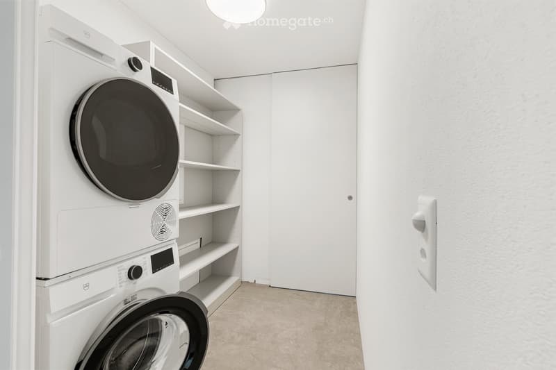 A modern, well-lit laundry room with a stacked washer and dryer unit, white shelves, and a neutral color palette.
