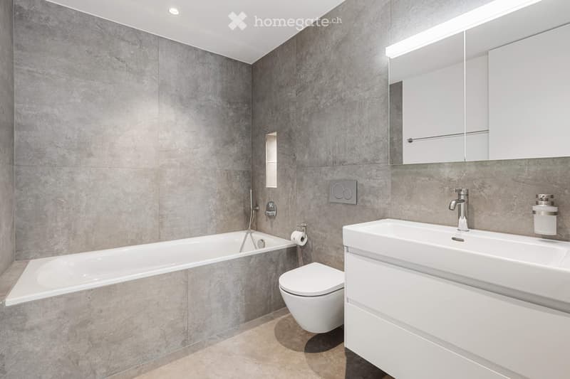 Modern bathroom with grey tiled walls, a bathtub, toilet, sink, and a mirrored cabinet.