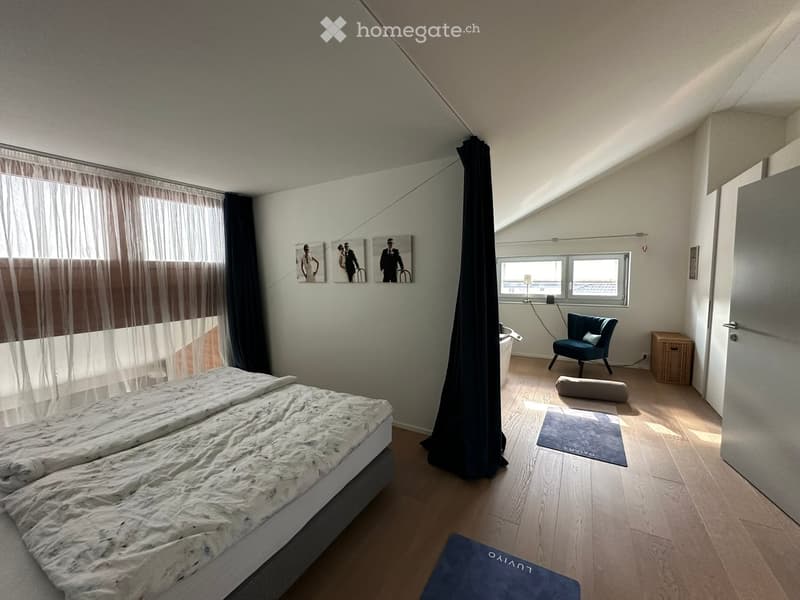 Bright bedroom with wooden flooring, large bed, blue armchair, picture frames on wall, and sheer curtains on windows.