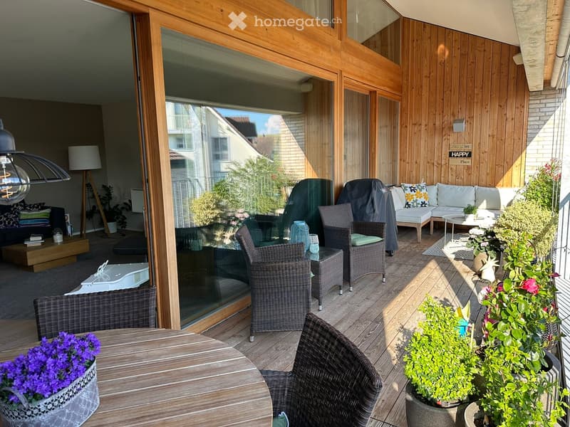 Balcony with wooden decking, outdoor furniture including a sofa, table, and chairs, surrounded by potted plants and flowers.
