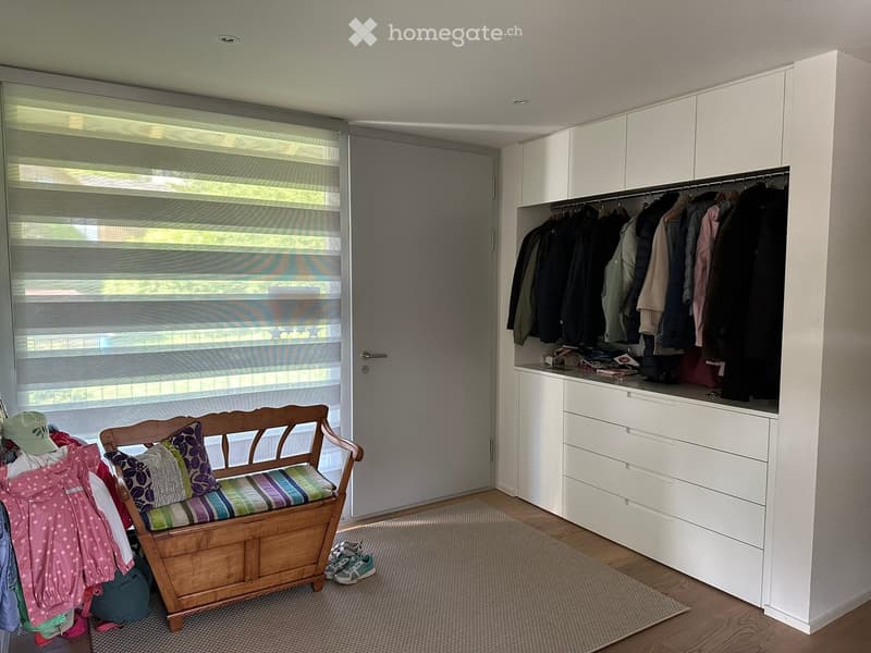 A well-lit hallway with a wooden bench, colorful pillows, a white wardrobe with hanging clothes and drawers, and a few personal items like a hat and shoes.