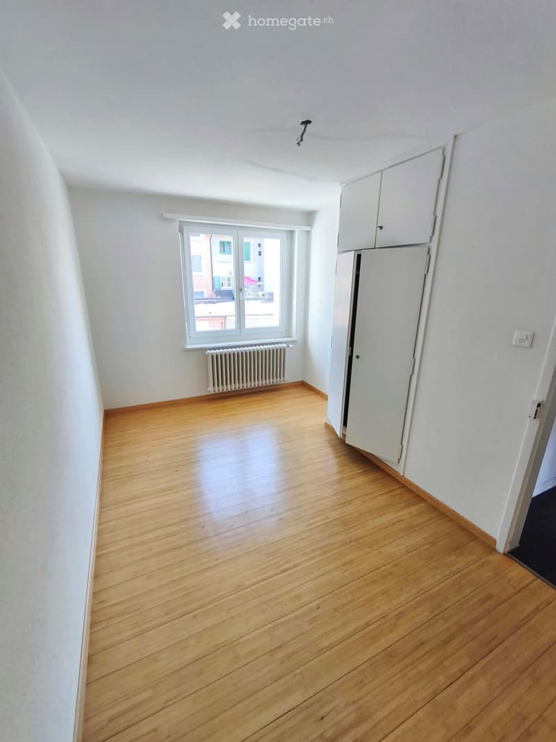 Empty room with wooden flooring, white walls, a window with a radiator below, and a partially visible cabinet and refrigerator on the right side.