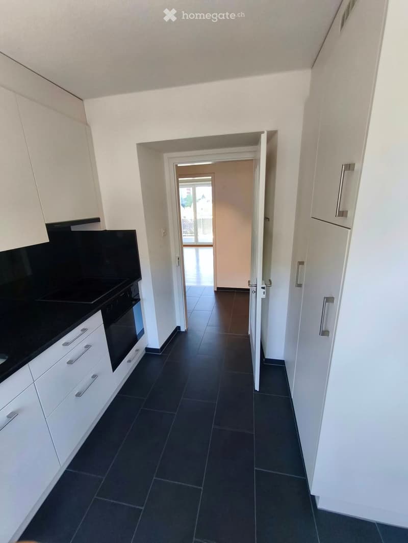 Modern hallway with white walls, dark tiled floor, and white cabinetry on one side. An open doorway leads to another room with visible furniture.