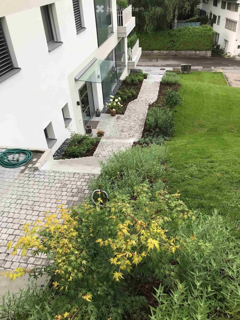 Garden area with a well-maintained lawn, various plants, shrubs, and a paved pathway leading to the house entrance. The house features large windows and a modern design.