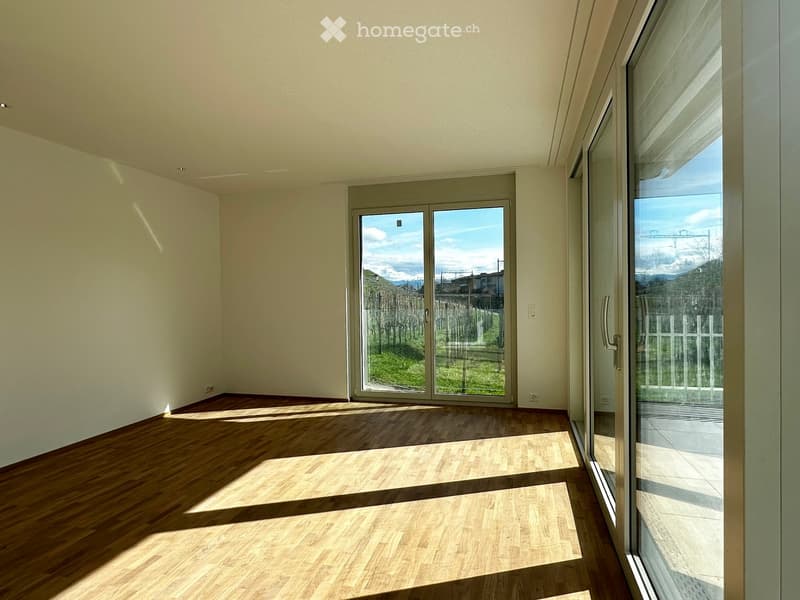 Empty room with wooden flooring and large glass doors leading to an outdoor area, featuring natural light and a view of greenery.