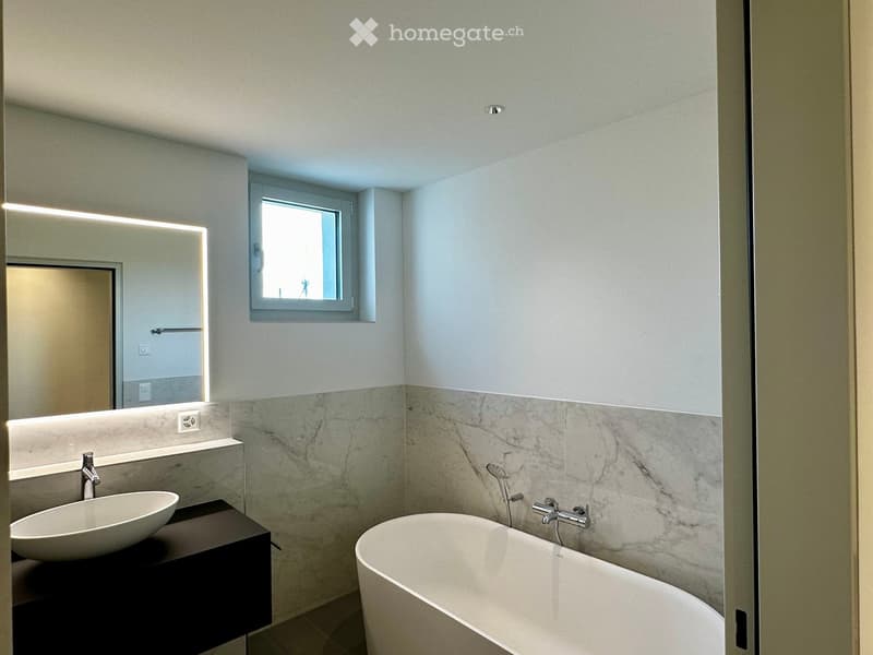 Modern bathroom with a white sink, bathtub, large mirror with LED lighting, and a window above the bathtub.