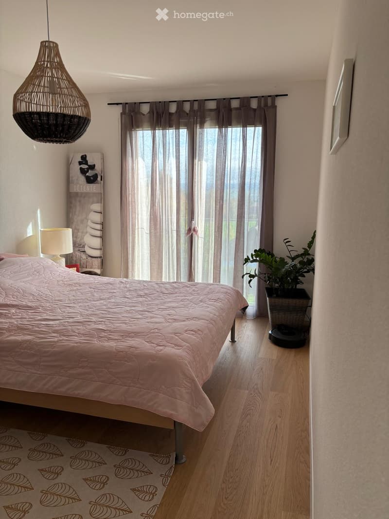 A well-lit bedroom with a wooden floor, a bed with a pink duvet, a table lamp, a wicker pendant light, a large window with sheer curtains, a potted plant, and a decorative picture frame.