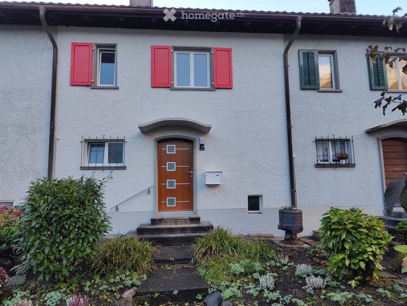 Two-story house with white exterior, red shutters, wooden front door, and well-maintained garden with various plants and shrubs.