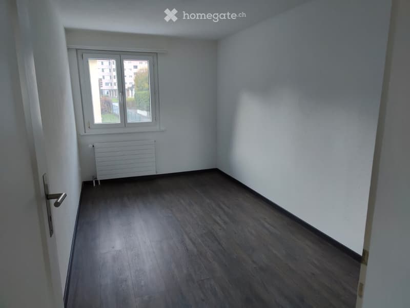 Empty room with wooden flooring, white walls, a window with white frame, and a radiator below the window.