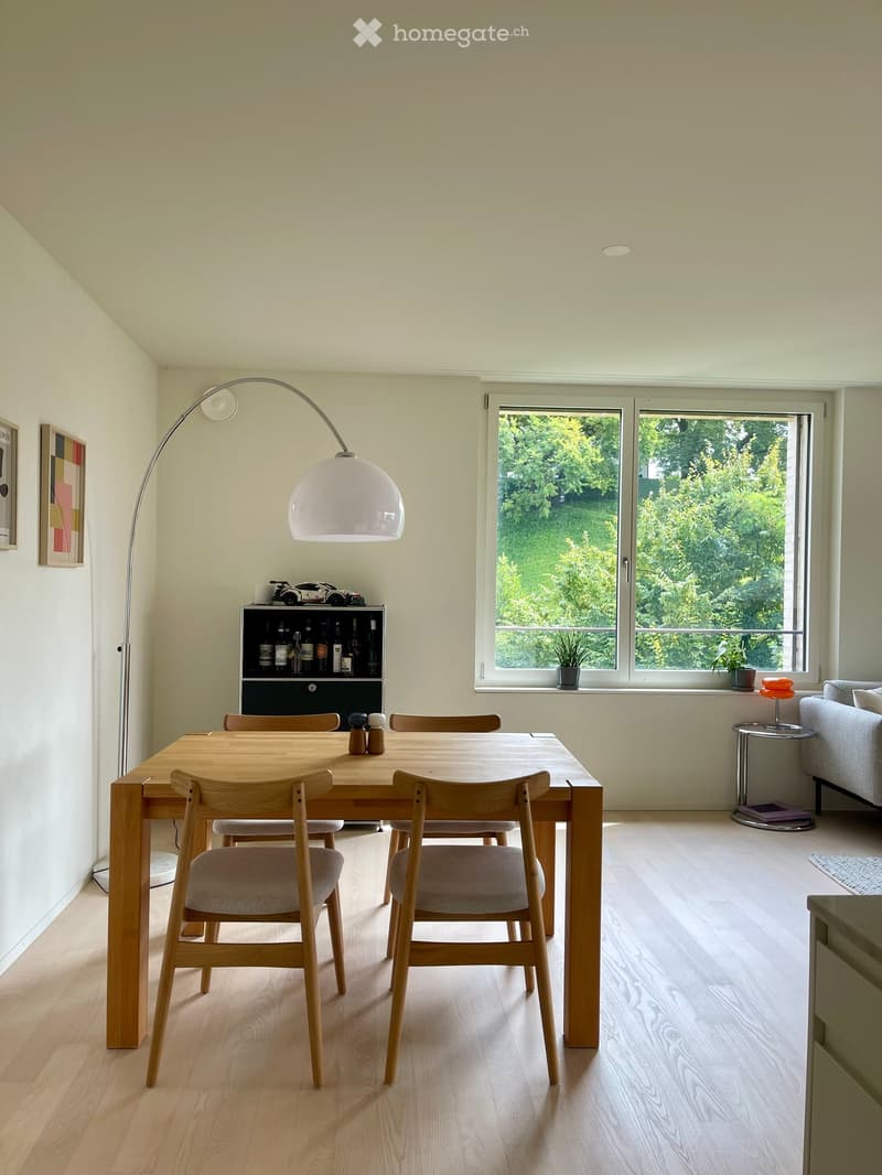 Dining room: wooden table, 4 wooden chairs, floor lamp, wooden cabinet with bottles, large window with green plants