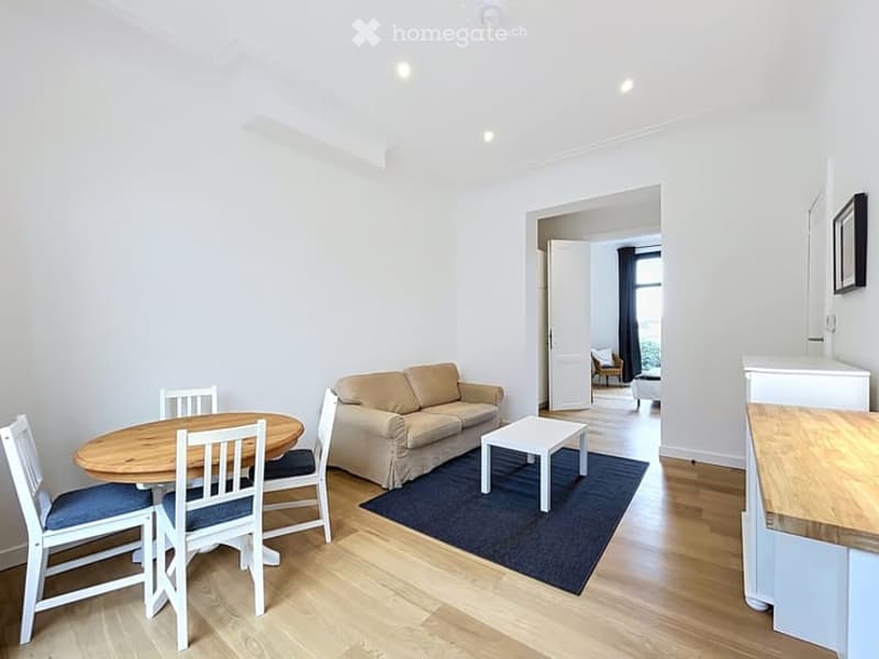 Bright, open-plan living and dining area with light wood flooring, white walls, and modern furnishings including a sofa, coffee table, dining table with chairs, and a kitchen counter.