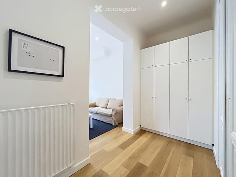 A modern hallway leading into a small living area with a cream sofa, coffee table, and white built-in wardrobes. The hallway features a radiator and a framed picture on the wall.