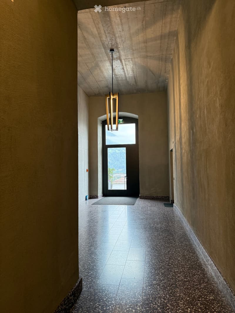 Modern hallway with polished tile flooring, textured walls, and a hanging light fixture. A glass door at the end leads to an outdoor view.