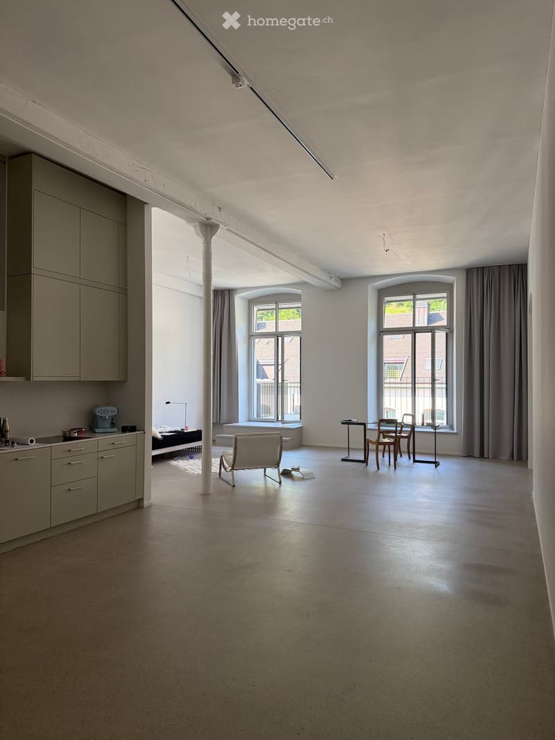 Open-plan room with high ceilings, large windows, and modern furniture including a chair, table, and sofa.