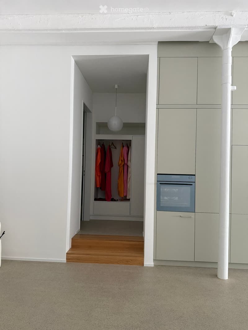 A modern, minimalist hallway leading to a small closet with a wardrobe and hanging clothes, and a built-in oven on the right side.