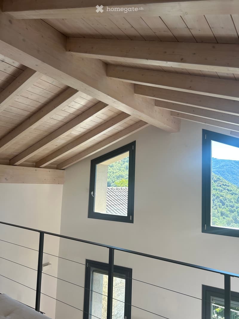 Modern hallway with exposed wooden ceiling beams, large windows offering scenic views, and sleek black metal railings.