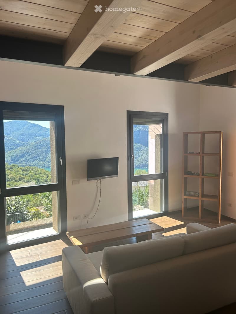 Modern living room with exposed wooden beams, large windows offering a scenic mountain view, a grey sofa, a wooden coffee table, a wooden shelf, and a wall-mounted television.