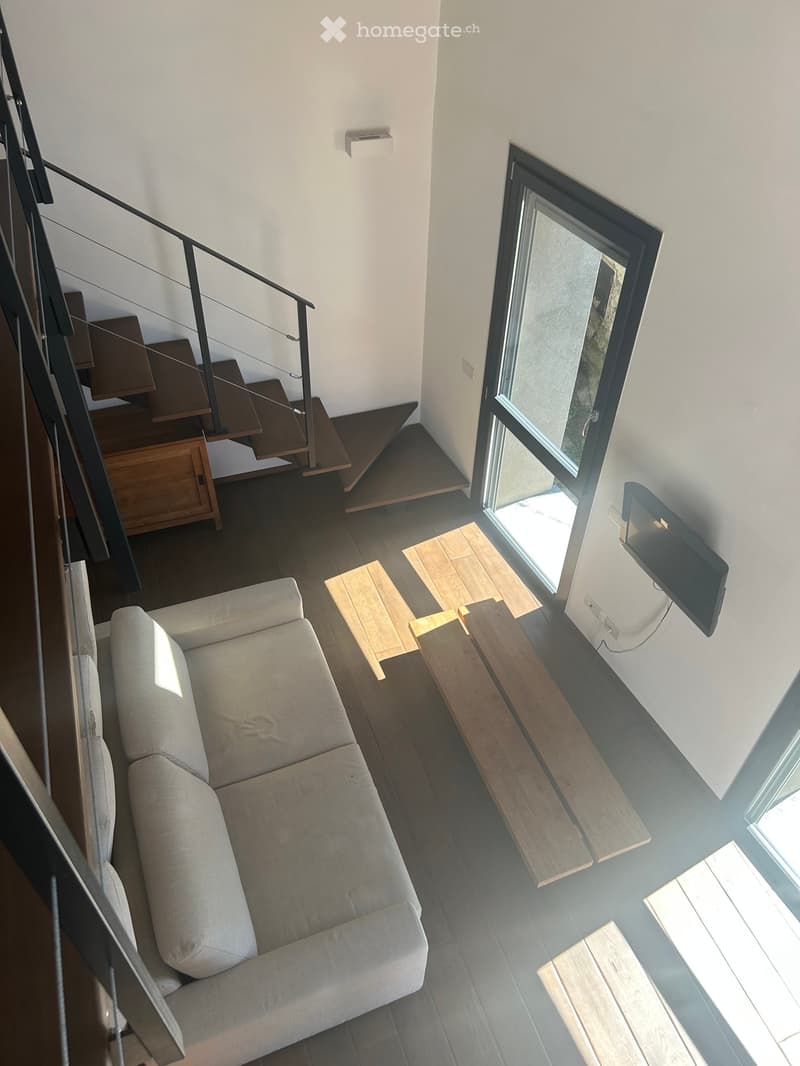 Modern living room with a grey sofa, coffee table, wooden stools, and a mounted screen. The room features a staircase leading to an upper level and large windows allowing natural light.