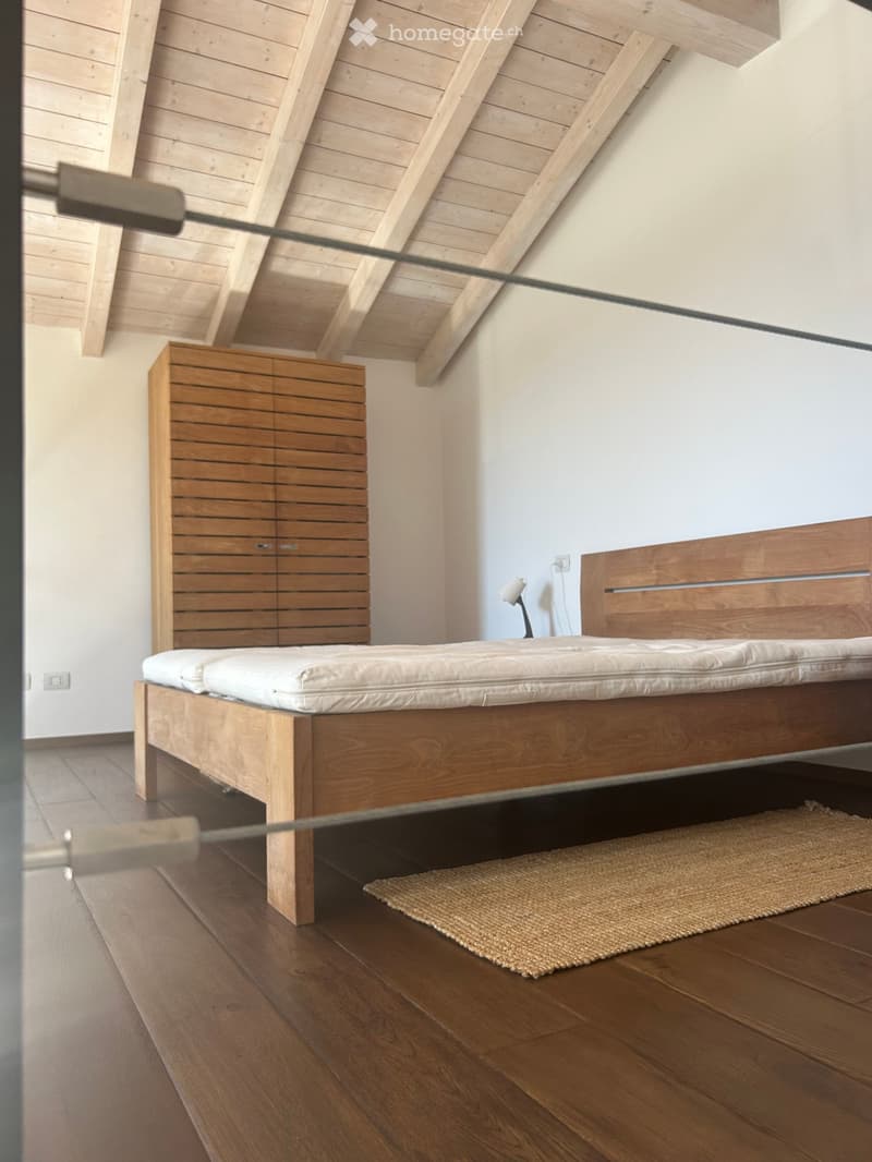 Modern bedroom with wooden bed frame, white mattress, nightstand, lamp, and small woven rug on wooden floor.