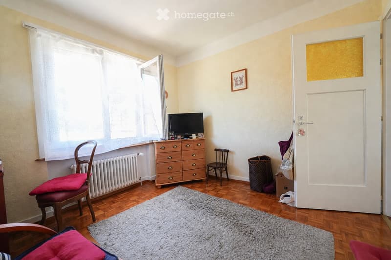 A furnished room with wooden flooring, a large window with white curtains, a television on a wooden cabinet, a wooden chair with red cushions, a small wooden stool, a gray shag rug, and a picture frame on the wall.