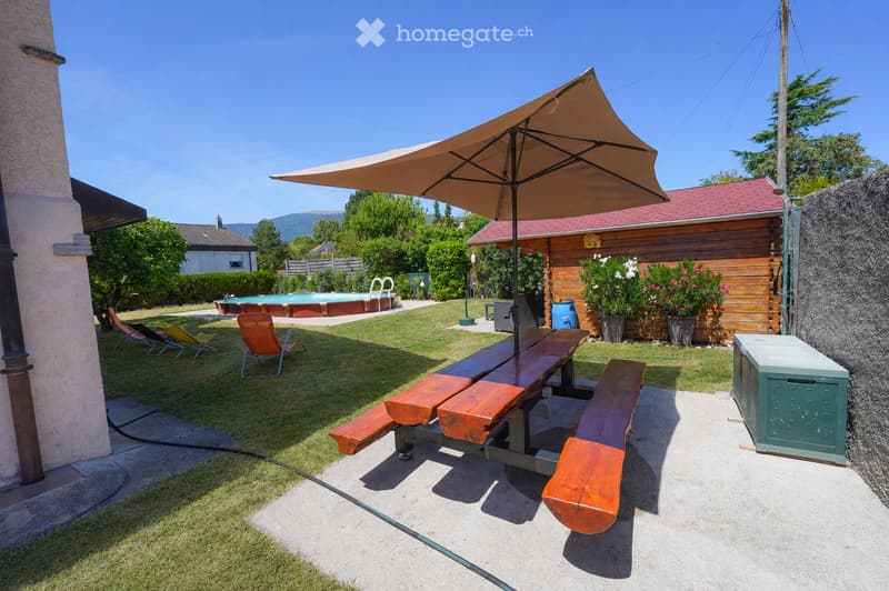 Garden with a wooden picnic table, benches, umbrella, lounge chairs, swimming pool, and surrounding greenery.