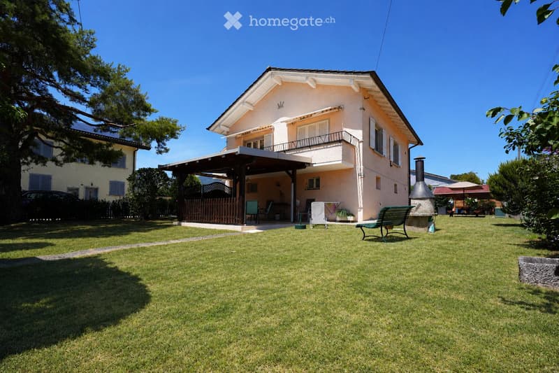 A well-maintained garden with a lawn, patio area, outdoor furniture, and a house with a balcony and covered porch.