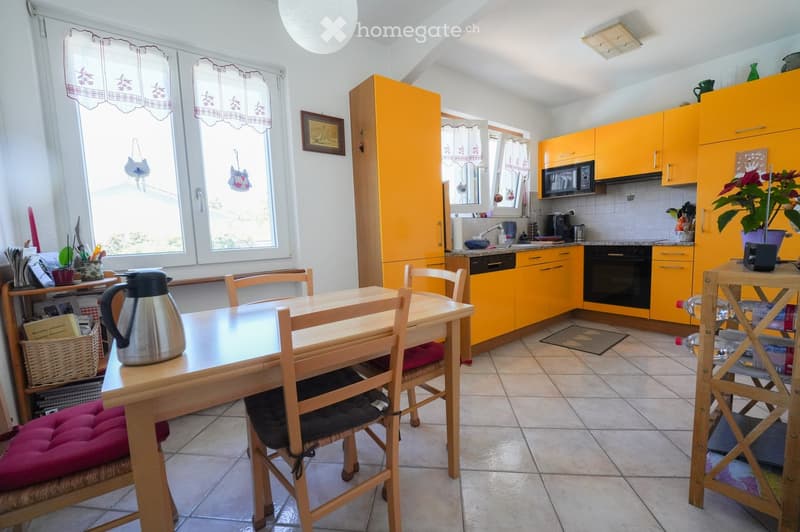 Bright kitchen with yellow cabinets, a wooden dining table with chairs, a microwave, oven, and various kitchen appliances. The space features large windows with curtains and a tiled floor.