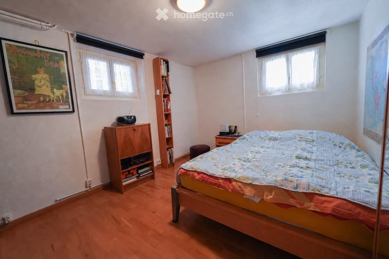 Cozy bedroom with wooden flooring, a bed with a colorful quilt, a nightstand, a small stool, a bookcase, a cabinet, and a vintage-style radio on top of the cabinet.