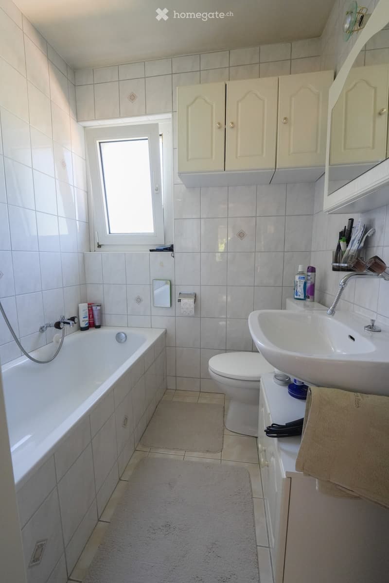 Bathroom with white tiles, bathtub, toilet, sink, mirror, and cabinet. Features a small window and a towel on the sink counter.