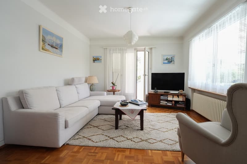 Bright living room with white walls, wooden flooring, and large windows. Features a grey sectional sofa, a coffee table with a patterned runner, an armchair, a table lamp, a television on a wooden stand, and several picture frames on the walls.