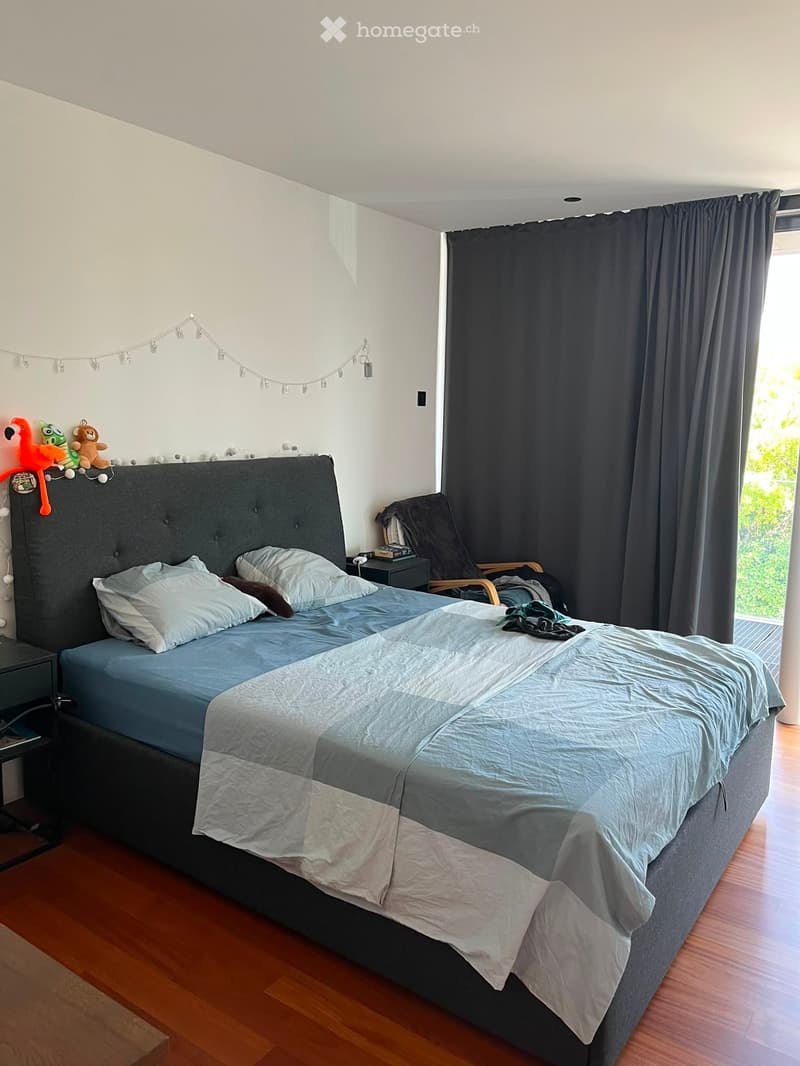 A modern bedroom with a large bed, nightstand, and a chair near the window. The room features wooden flooring, white walls, and dark curtains.
