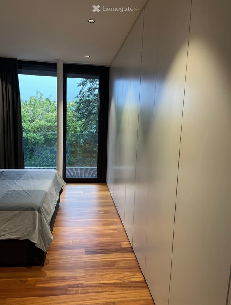 Modern bedroom with wooden flooring, large windows, and a bed with a grey duvet.