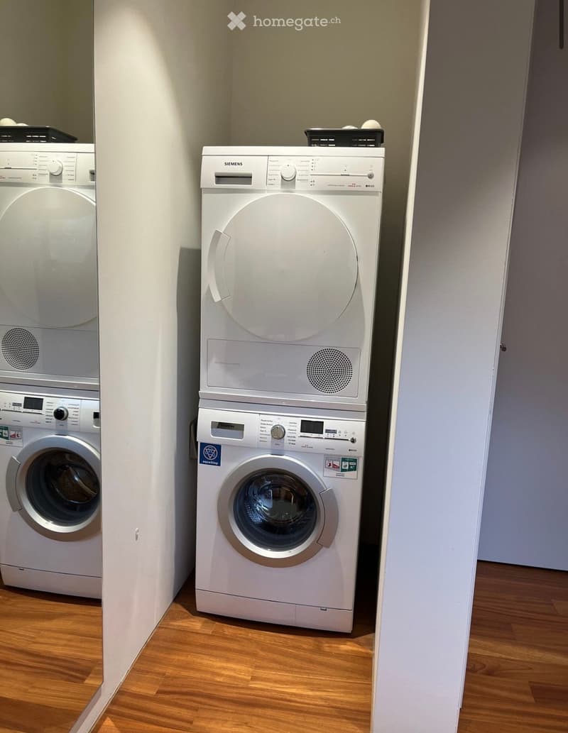 A small room with wooden flooring and two white appliances: a washing machine and a dryer, both with control panels and dials.