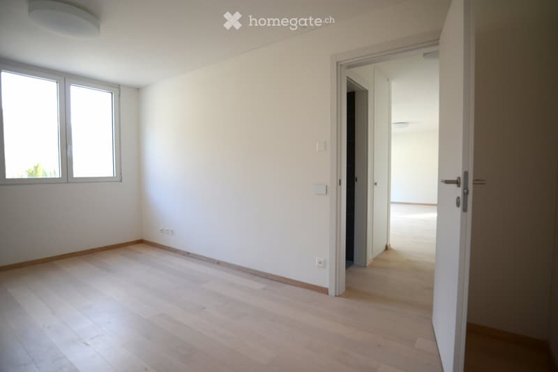 Empty room with light wooden flooring, white walls, and large windows allowing natural light. The room features a door leading to another space.