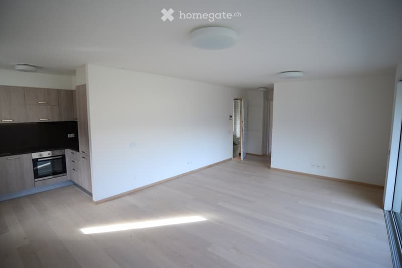 Empty room with light wooden flooring, white walls, and a small kitchen area with cabinets and an oven. The space is well-lit with recessed lighting.