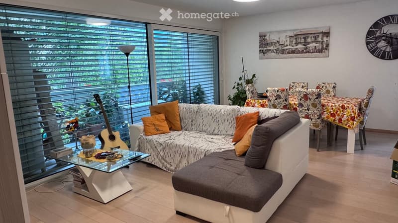Modern living room with large windows, white sofa with patterned cover, glass coffee table, dining area with floral tablecloth, and a guitar near the window.