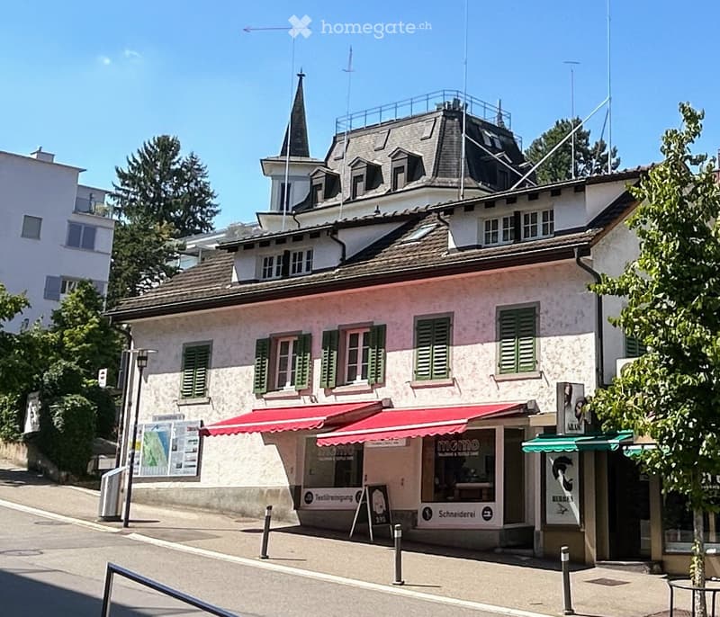 Two-story building with a red awning, green shutters, and a spire on the roof. The ground floor has storefronts with signage for 'momo' and 'Schneiderei'.