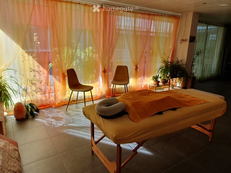 A serene massage room with a massage table, two chairs, a small stool, and potted plants, illuminated by natural light through sheer orange curtains.